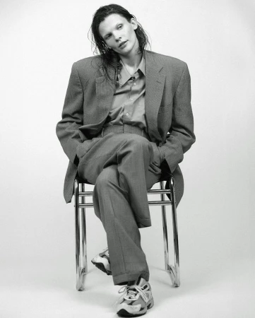 Model with wet hair in an oversized grey suit sitting on a chair against a white seamless background