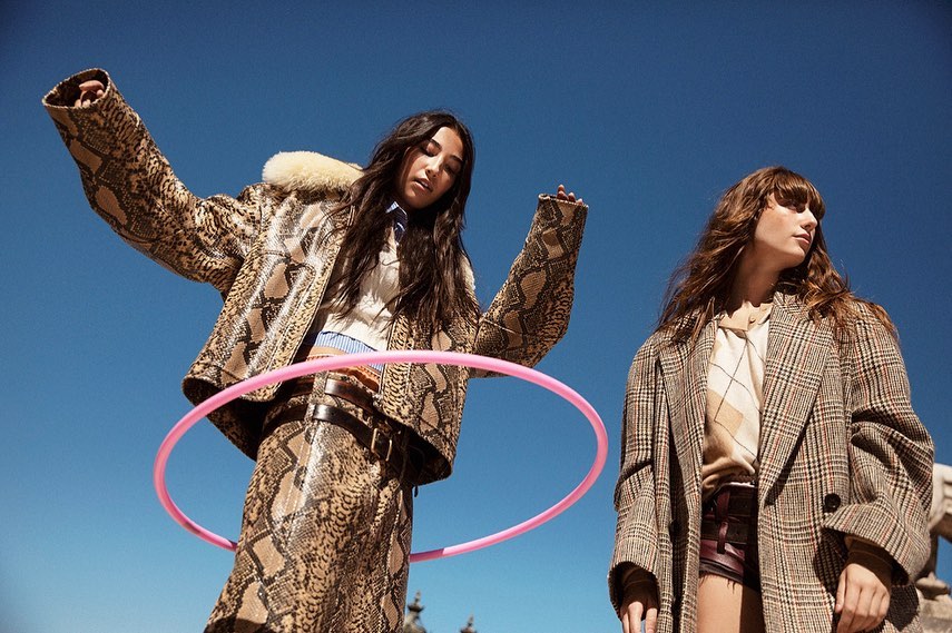 Two women in patterned coats pose with a pink hula hoop against a blue sky backdrop. Fashionable outerwear is the focus.