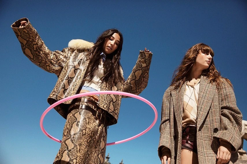 Two women in patterned coats pose with a pink hula hoop against a blue sky backdrop. Fashionable outerwear is the focus.
