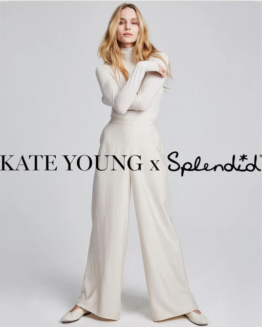 A blonde adult woman poses in a studio wearing a long-sleeved light-beige top and flowing light-beige pants with off-white shoes.