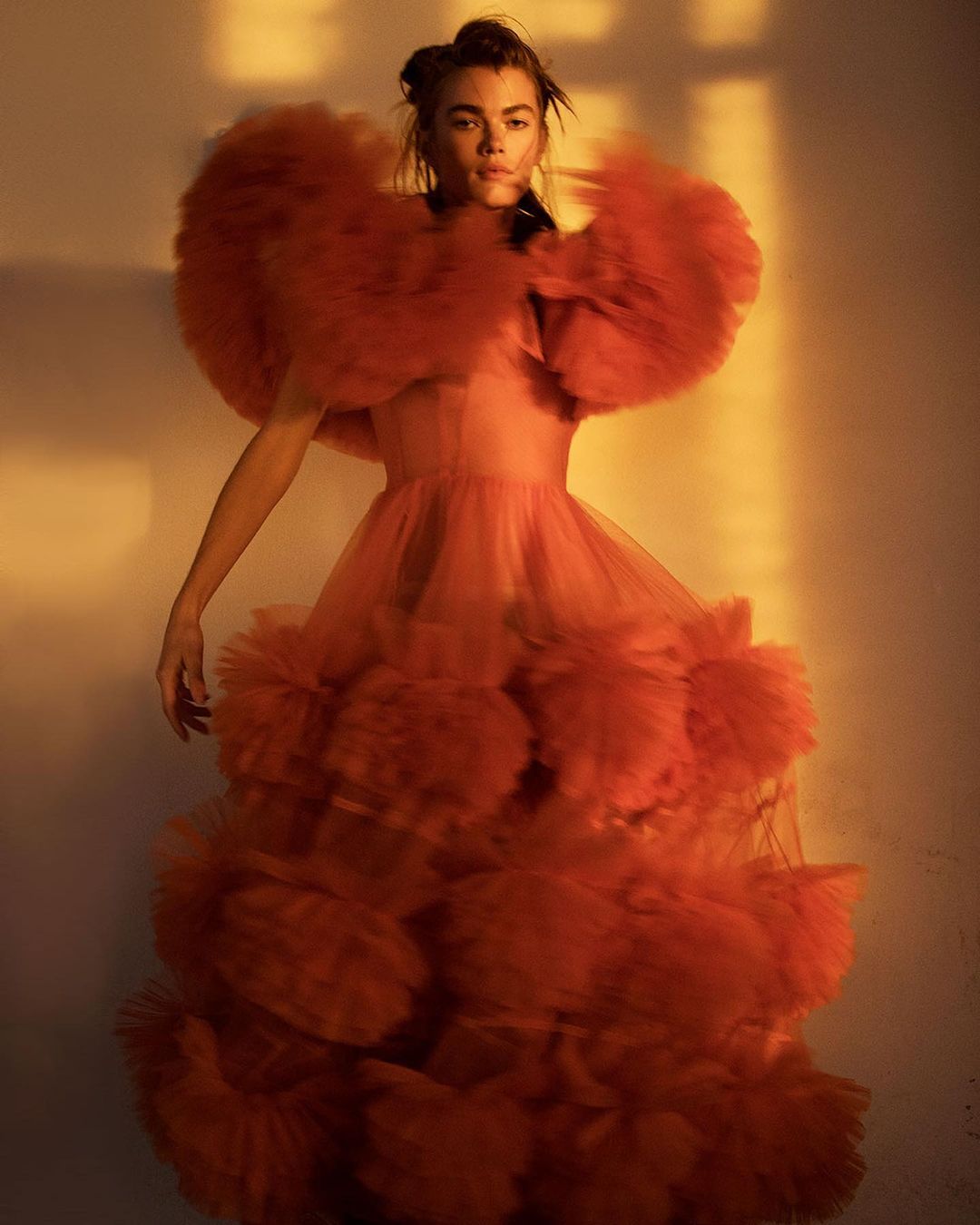 A woman in a red ruffled gown with large ruffled sleeves poses in a photo studio with natural light, showcasing a high-fashion editorial style.