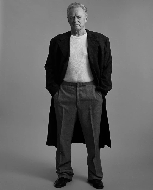 Full body studio portrait of adult man in a dark coat, white shirt, and grey pants, hands in pockets, standing against grey backdrop.