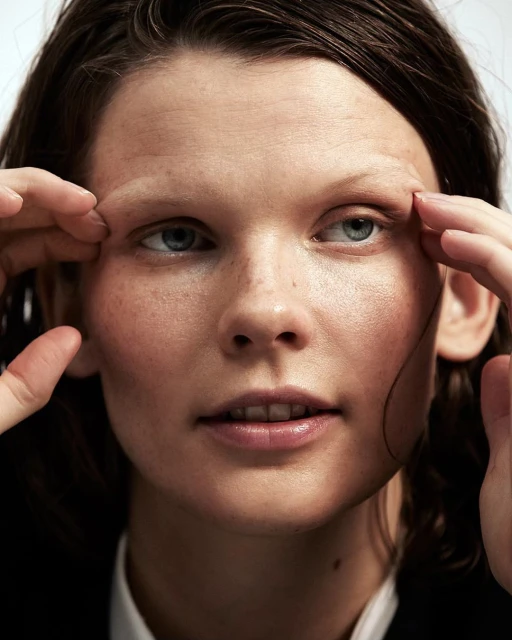Close-up portrait of a woman with fair skin and wet hair, touching her face with both hands, set against a bright white background.