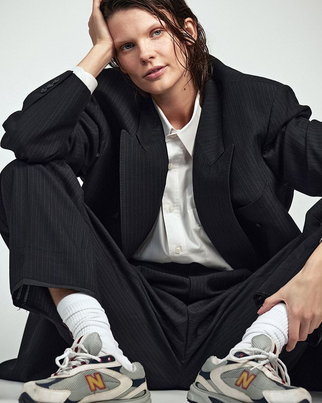 Stylish woman in pinstripe suit and athletic shoes sits in a photo studio, casually posing with wet hair and a confident expression.