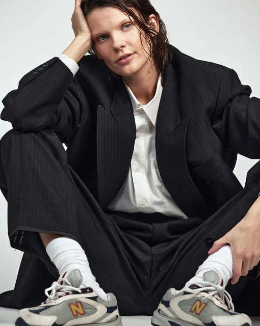 Stylish woman in pinstripe suit and athletic shoes sits in a photo studio, casually posing with wet hair and a confident expression.