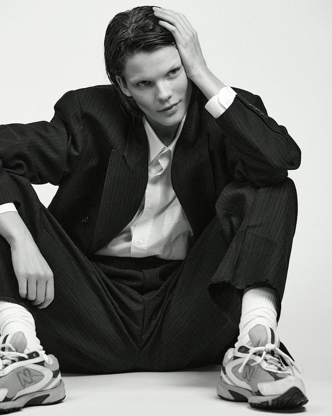 Monochrome portrait of a person in a pinstripe suit, white shirt, socks and sneakers, seated against a plain backdrop in a studio setting.