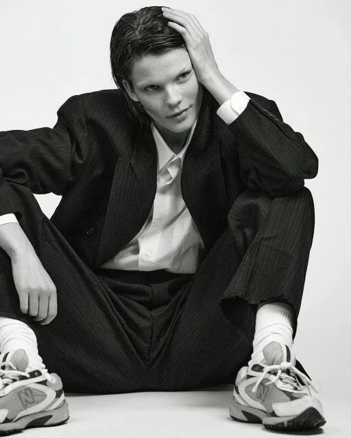 Monochrome portrait of a person in a pinstripe suit, white shirt, socks and sneakers, seated against a plain backdrop in a studio setting.