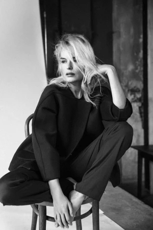 Black and white portrait of a blonde woman sitting on a wooden chair wearing a dark blazer and trousers.