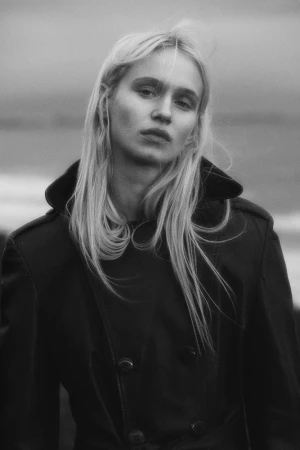 Monochrome portrait of a blonde woman wearing a dark double-breasted trench coat outdoors.