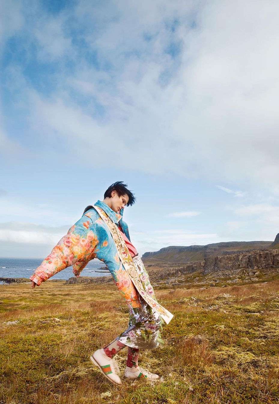 Model in vibrant floral print puffer jacket leaning backwards in a grassy meadow under a cloudy blue sky.