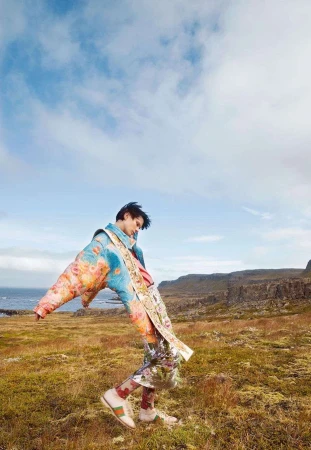 Model in vibrant floral print puffer jacket leaning backwards in a grassy meadow under a cloudy blue sky.