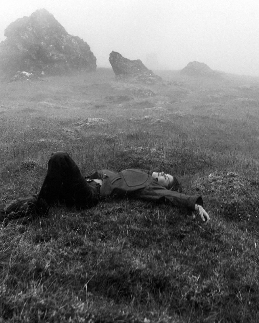 Black and white portrait of a person lying on grassy terrain in a misty landscape with large rock formations