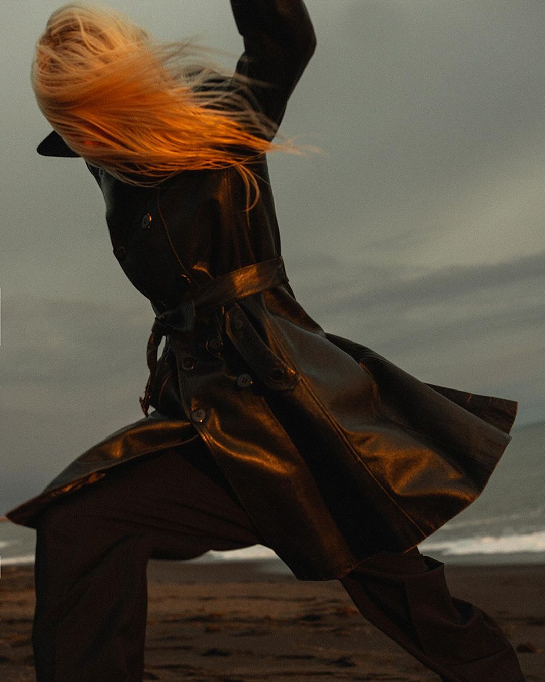 Model with long blonde hair in a black leather coat moving dynamically on a sandy beach during sunset.