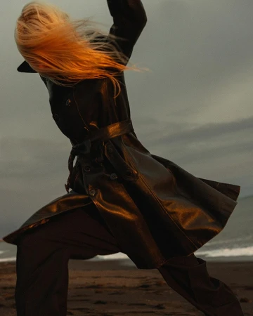 Model with long blonde hair in a black leather coat moving dynamically on a sandy beach during sunset.