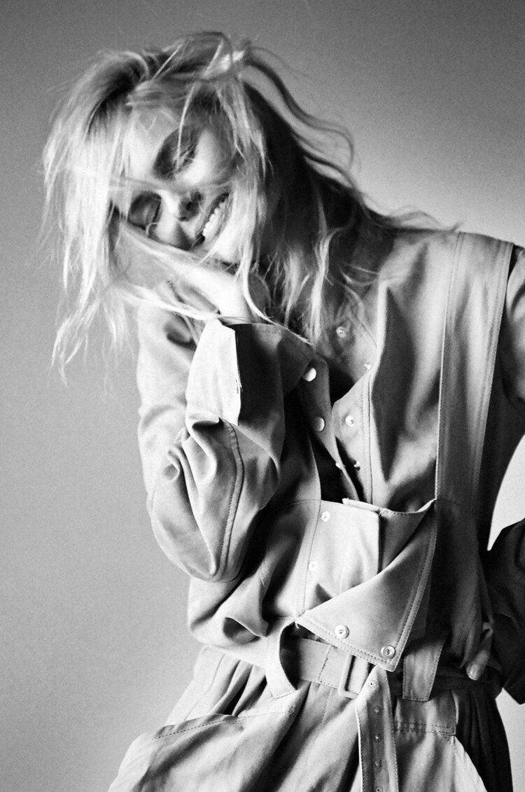 Black-and-white portrait of a smiling blonde woman in a button-down jumpsuit, captured in a naturally lit indoor setting.