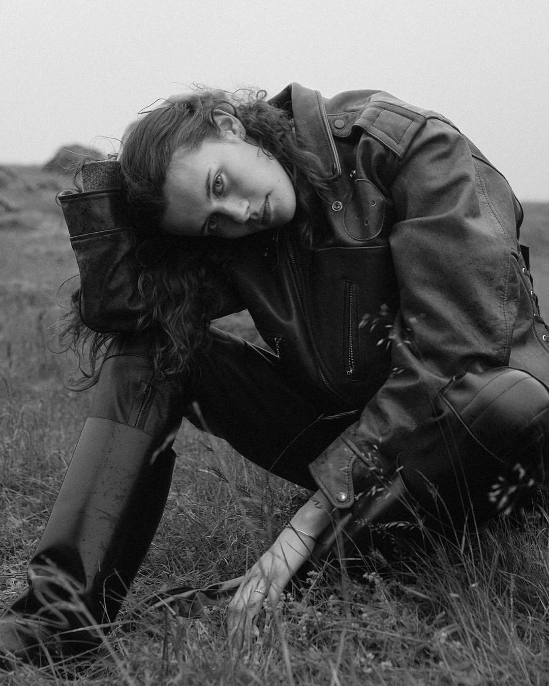 Black and white portrait of a woman with curly hair wearing a leather jacket, sitting in a grassy field. Moody outdoor fashion shot.