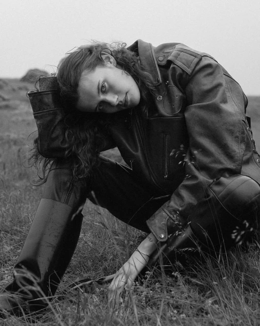 Black and white portrait of a woman with curly hair wearing a leather jacket, sitting in a grassy field. Moody outdoor fashion shot.