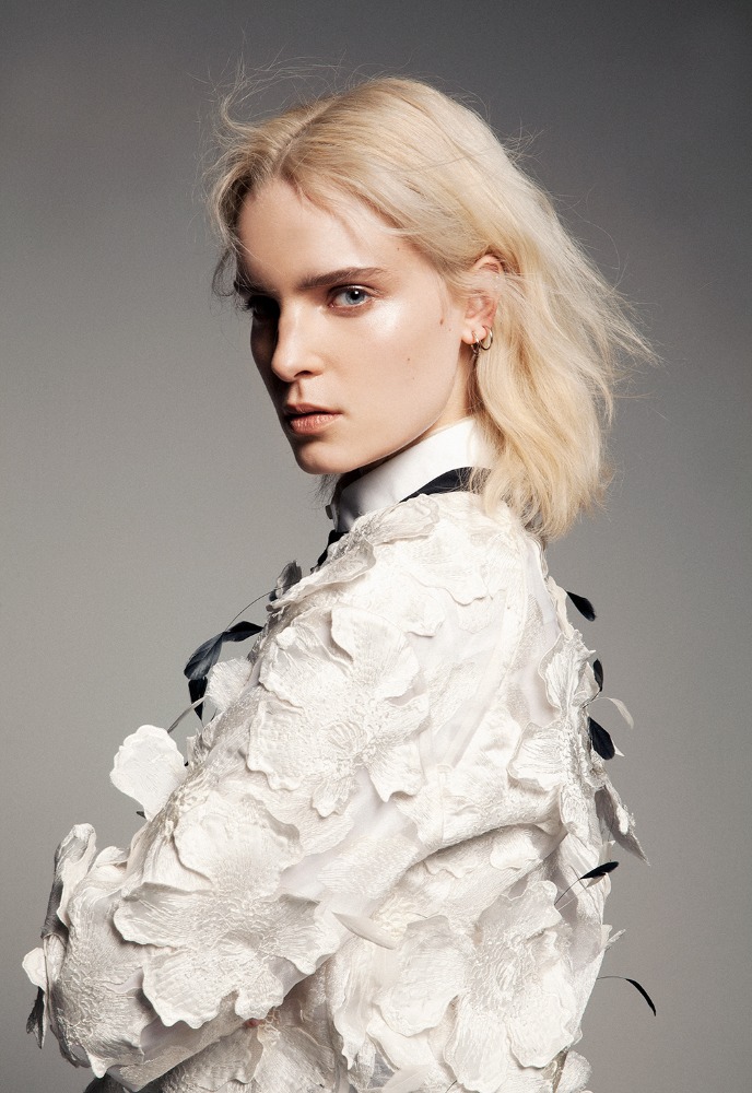 Blonde woman wearing white floral textured garment looking back over her shoulder against a neutral grey background