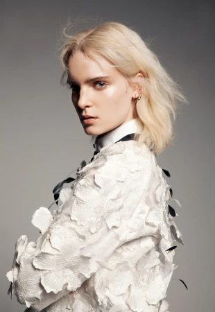 Blonde woman wearing white floral textured garment looking back over her shoulder against a neutral grey background