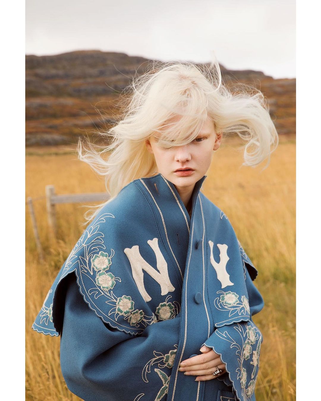 Blonde woman posing in an open field wearing an embroidered blue shawl with 'NY' initials, mountains in the background and cloudy sky.