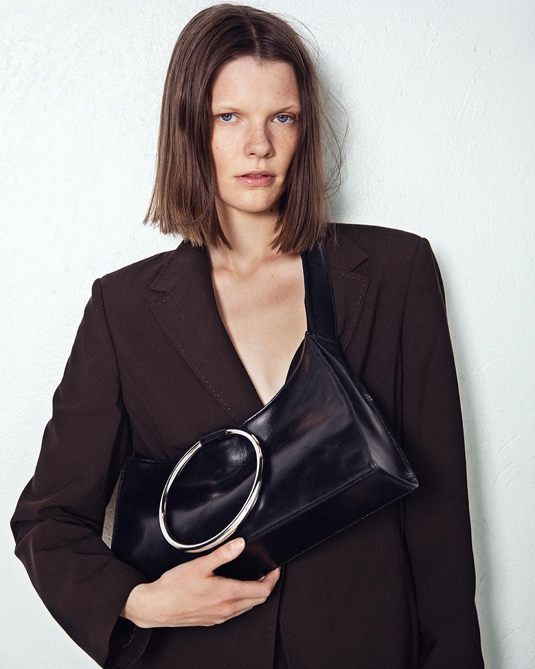 Model in brown blazer holding a black leather handbag with a large silver ring detail against a white wall