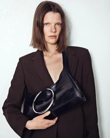 Model in brown blazer holding a black leather handbag with a large silver ring detail against a white wall