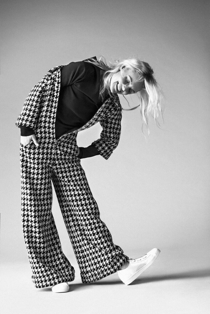 Woman in houndstooth suit strikes a pose in photo studio. She's bending over with a slight smile and white shoes, on neutral backdrop.