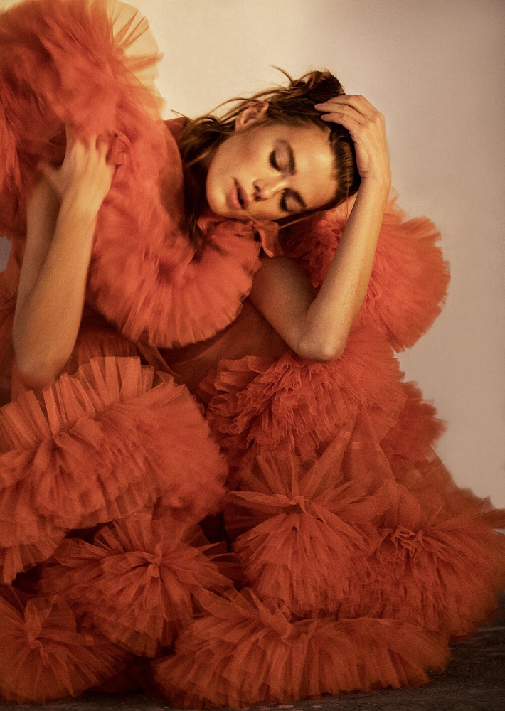 A woman wearing a large orange ruffled gown in a studio setting, eyes closed, with her hand resting on her head.