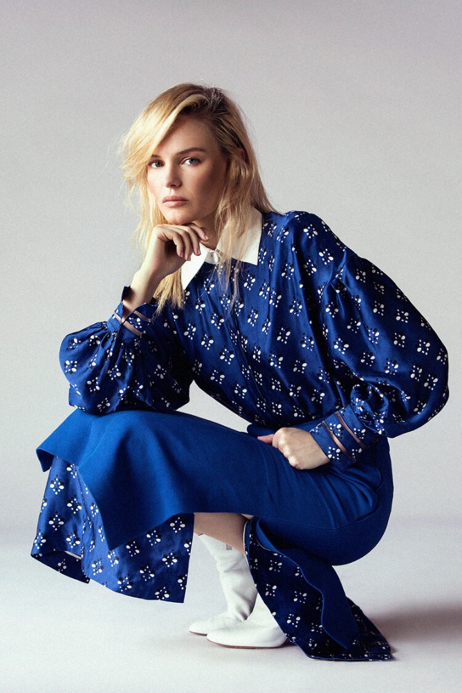 A blonde-haired adult woman in a blue patterned blouse and skirt is crouched and posed in a studio setting, with soft lighting and a clean background.