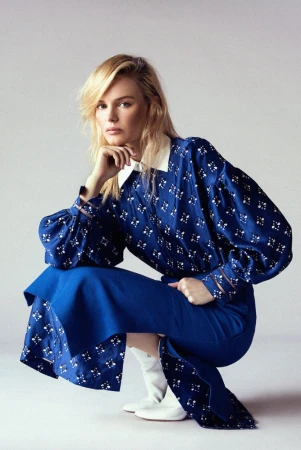 A blonde-haired adult woman in a blue patterned blouse and skirt is crouched and posed in a studio setting, with soft lighting and a clean background.