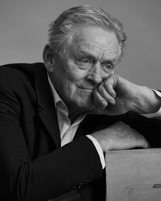 Black and white portrait of a mature adult man in a dark suit with a white shirt, resting his face on his hands with a thoughtful expression.