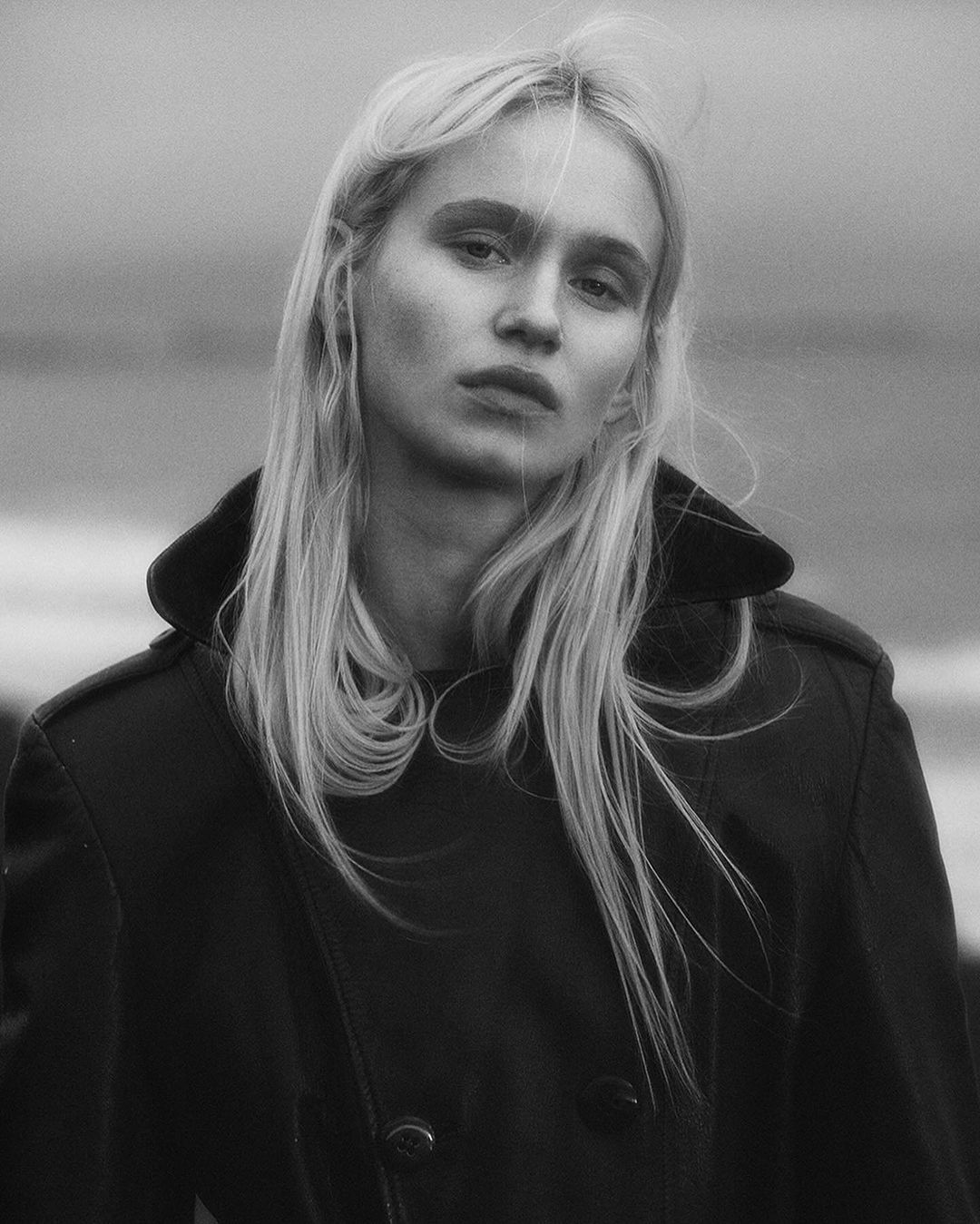 Black and white portrait of a blond adult woman wearing a black overcoat, set against an outdoor background with muted tones.