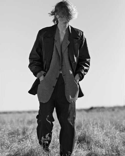 Black and white portrait of a person in a patterned suit and leather jacket standing in a grassy field under a clear sky.