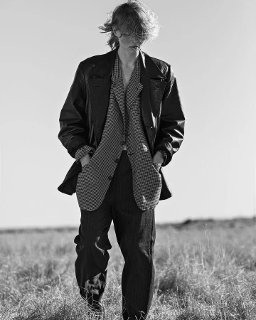Black and white portrait of a person in a patterned suit and leather jacket standing in a grassy field under a clear sky.