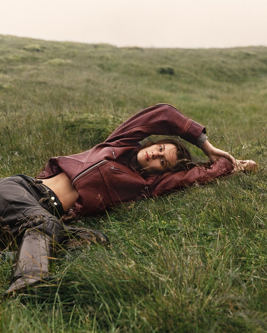 Woman in burgundy jacket lying in green field; brunette woman relaxing in grassy landscape with leather boots and casual fashion apparel.