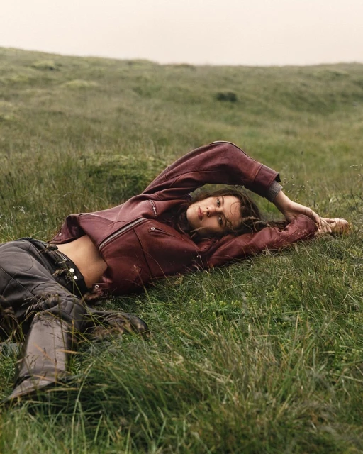 Woman in burgundy jacket lying in green field; brunette woman relaxing in grassy landscape with leather boots and casual fashion apparel.