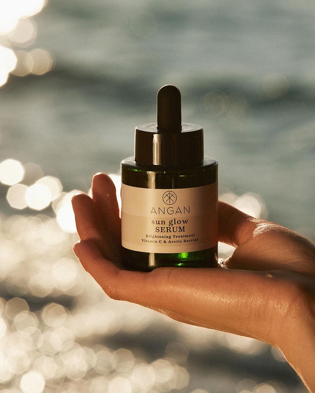 Close-up of a hand holding a dark green bottle of Argan Sun Glow Serum against a bokeh background of the sea and sunlight.