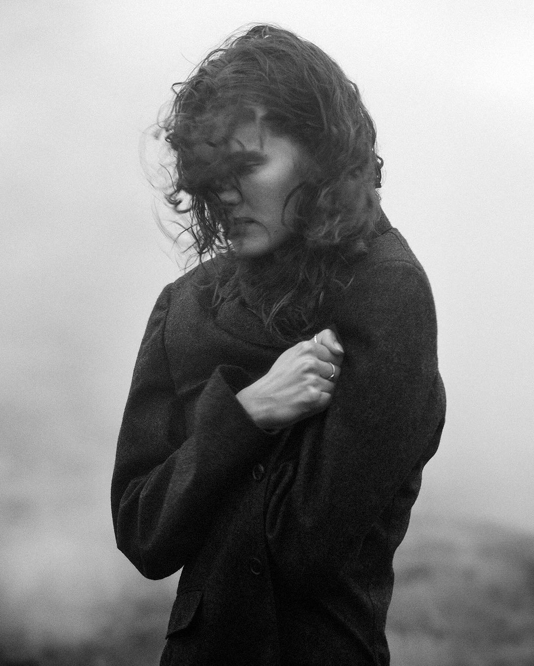 Monochrome portrait of an adult female with curly brown hair, wearing a dark coat, standing outdoors with a wind-swept hair effect.