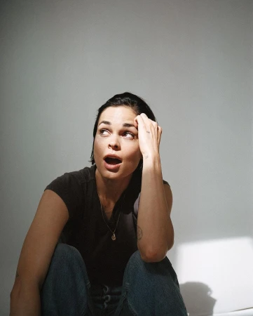 Woman in dark t-shirt with hand to head looking up against a plain grey studio backdrop