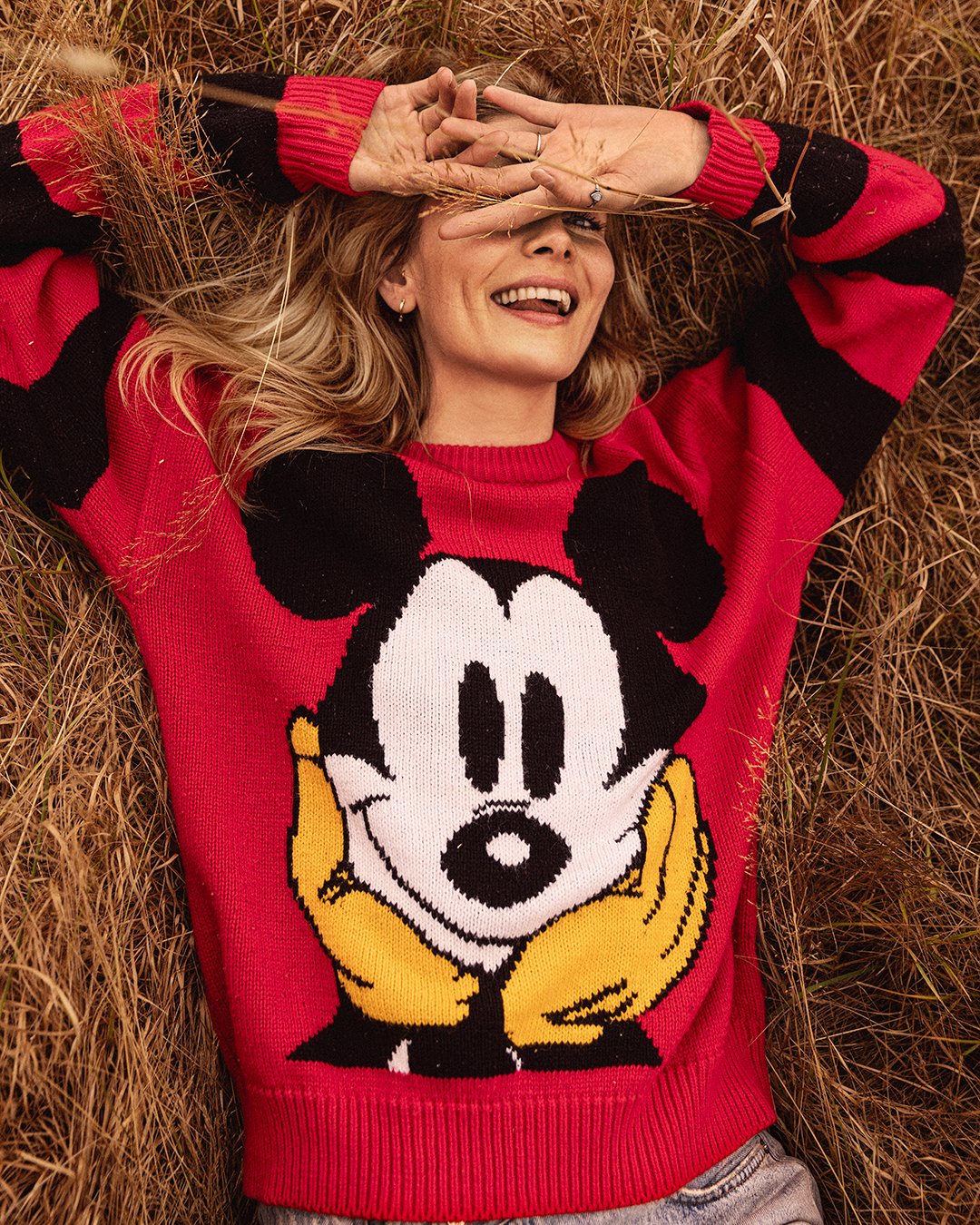 A woman with blonde hair is laying in tall brown grass wearing a red sweater with a Mickey Mouse graphic, and her hands above her face.