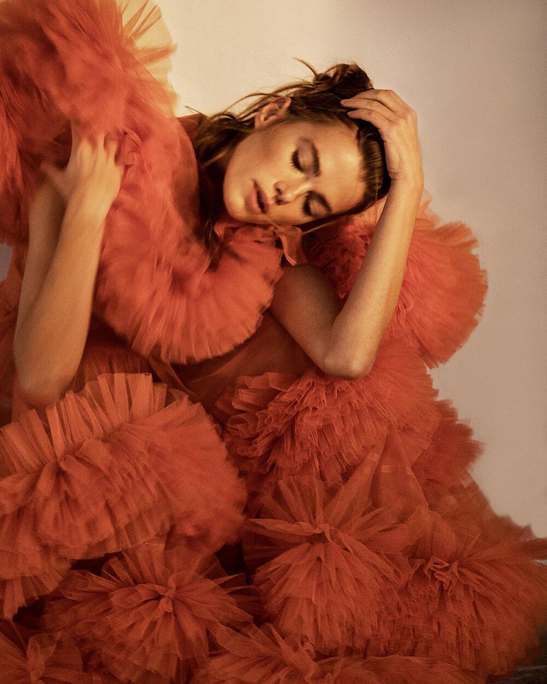 Adult female in a textured orange ruffled dress with hand on head posing indoors with soft light and medium shot in portrait orientation.