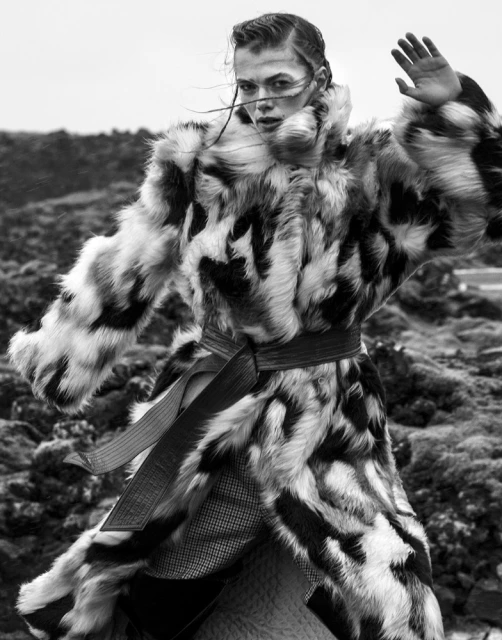Monochrome portrait of a woman in a patterned fur coat with a belt, posing outdoors against a rocky landscape under overcast skies.