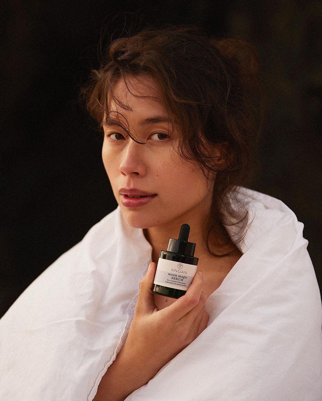 Adult woman wrapped in a white blanket holds a black bottle of beauty serum. Natural beauty, skincare product, minimal composition.
