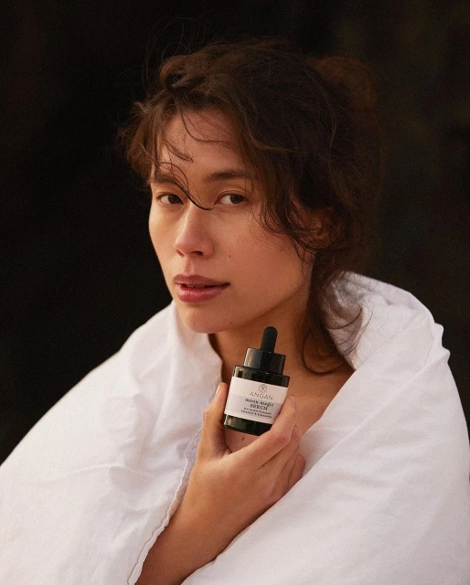 Adult woman wrapped in a white blanket holds a black bottle of beauty serum. Natural beauty, skincare product, minimal composition.