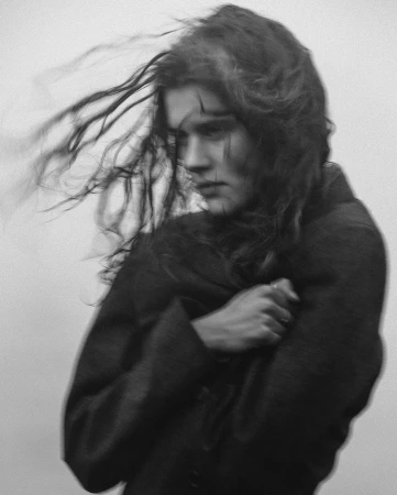 Monochrome portrait of a woman with dark, wavy hair, wearing a coat, with a blurred aesthetic and neutral studio background, in a wind effect.