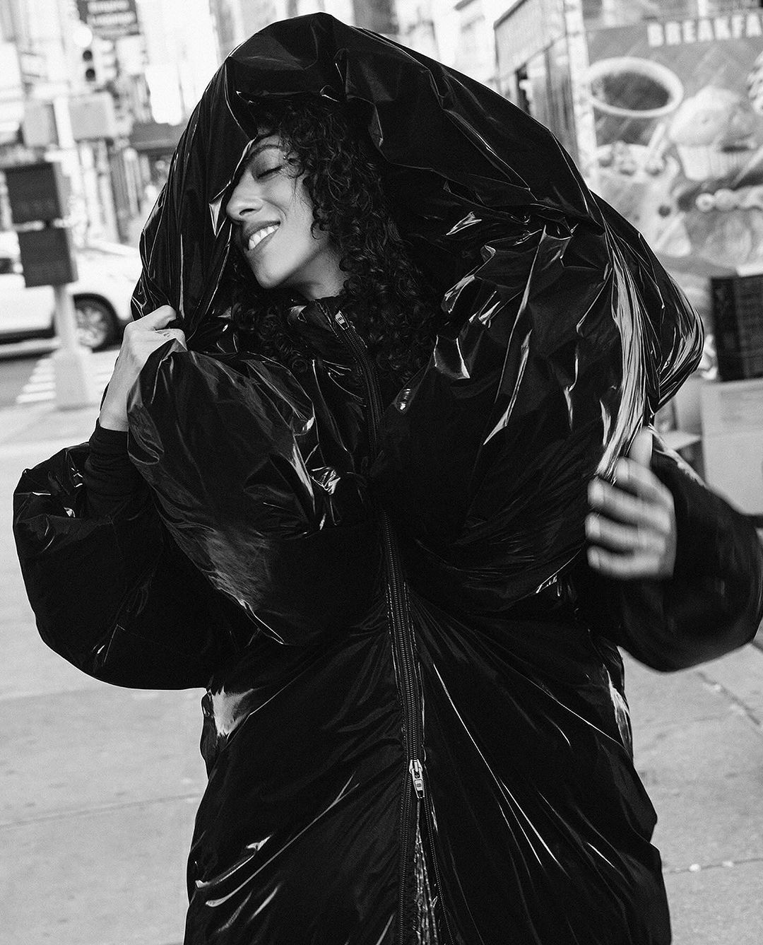 Fashion editorial shot of a woman wearing an oversized, glossy black puffer coat with the hood up, posing on a city street.