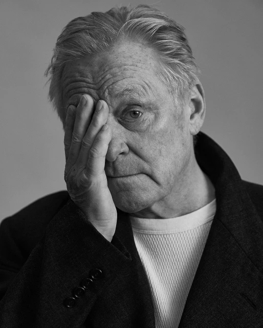 Black-and-white portrait of an elderly man with hand on face, wearing a coat and a white undershirt, studio shot.