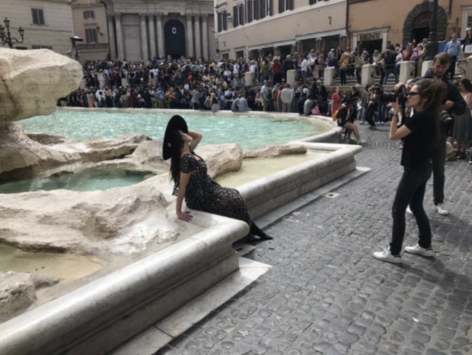Woman poses by the Trevi Fountain in Rome, Italy, while a photographer takes her picture. A crowd gathers in the background.