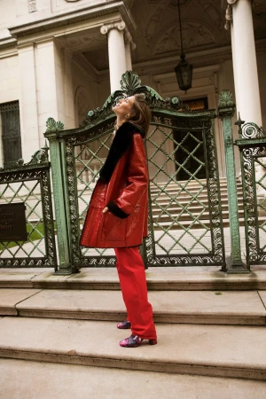 Woman in a red patent leather coat and pants stands on stone steps outside a building with iron gates, looking up, in a fashion shot.