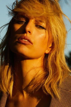 Close-up portrait of a blonde adult female in warm lighting wearing a tan blazer against a blurred neutral background.